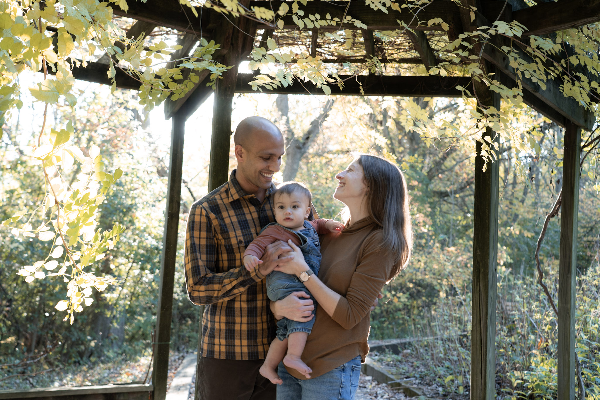 Parents hold their child beneath a wooden pergola covered in vines